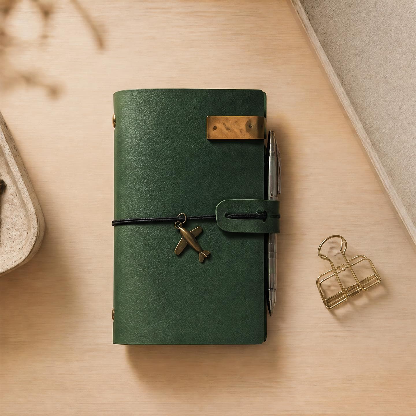 Dark emerald green leather travel journal with a brass clip and airplane charm on light wood