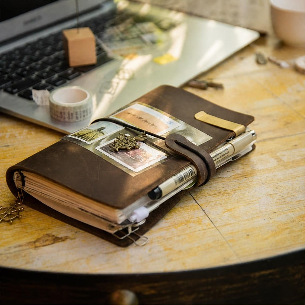 Dark brown leather travel journal on a wooden desk with stamps and a ship's wheel charm